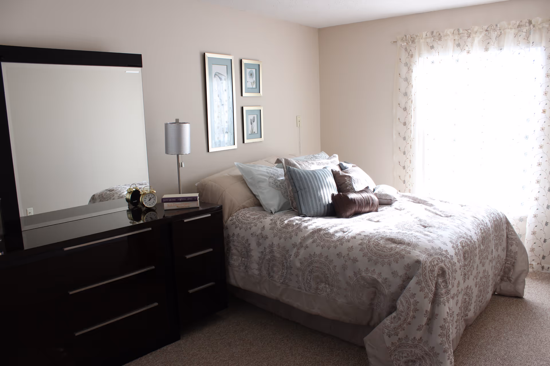 A bedroom with a neatly made bed featuring multiple pillows and a patterned bedspread. Next to the bed is a dark wooden dresser with a large mirror, a table lamp, an alarm clock, and a book. The room has light-colored walls and a window with sheer floral curtains letting in natural light.