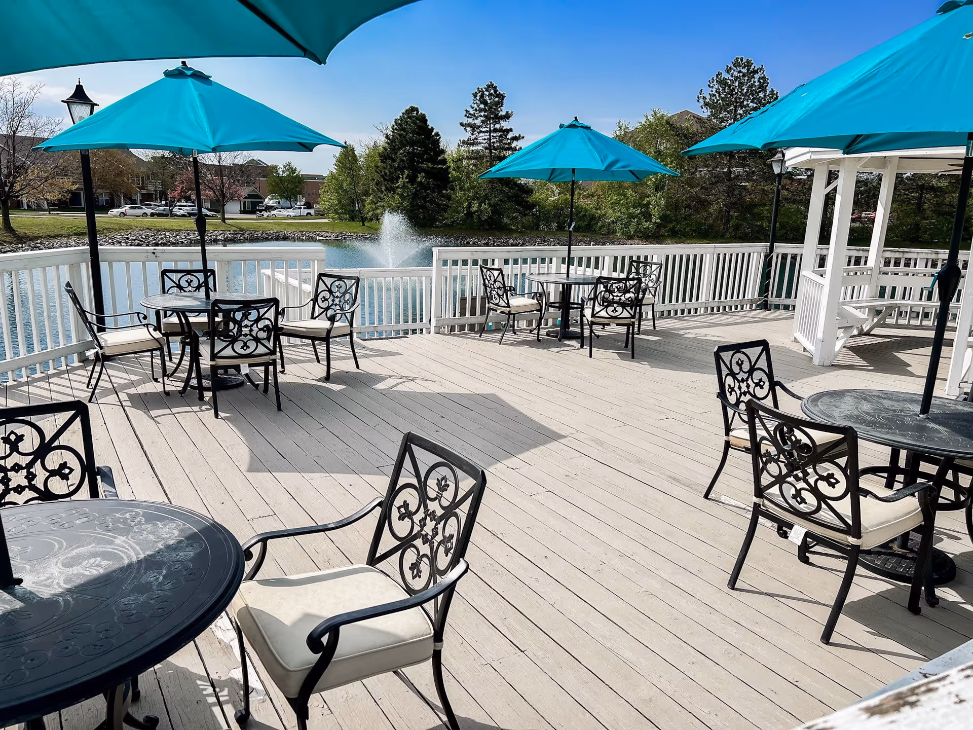 Outdoor deck area with several black metal tables and chairs with white cushions, shaded by turquoise umbrellas. The deck overlooks a pond with a water fountain, surrounded by trees and residential buildings in the background under a clear blue sky.