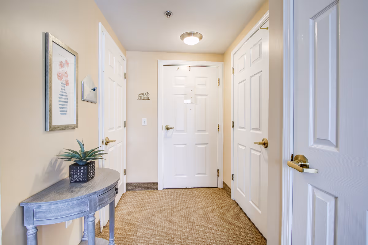A hallway area inside a senior living facility with beige walls and carpeted floor. There are three white doors with gold handles, a small gray wooden table with a potted plant on it, and a framed picture of flowers on the wall. A ceiling light fixture is visible above the central door.