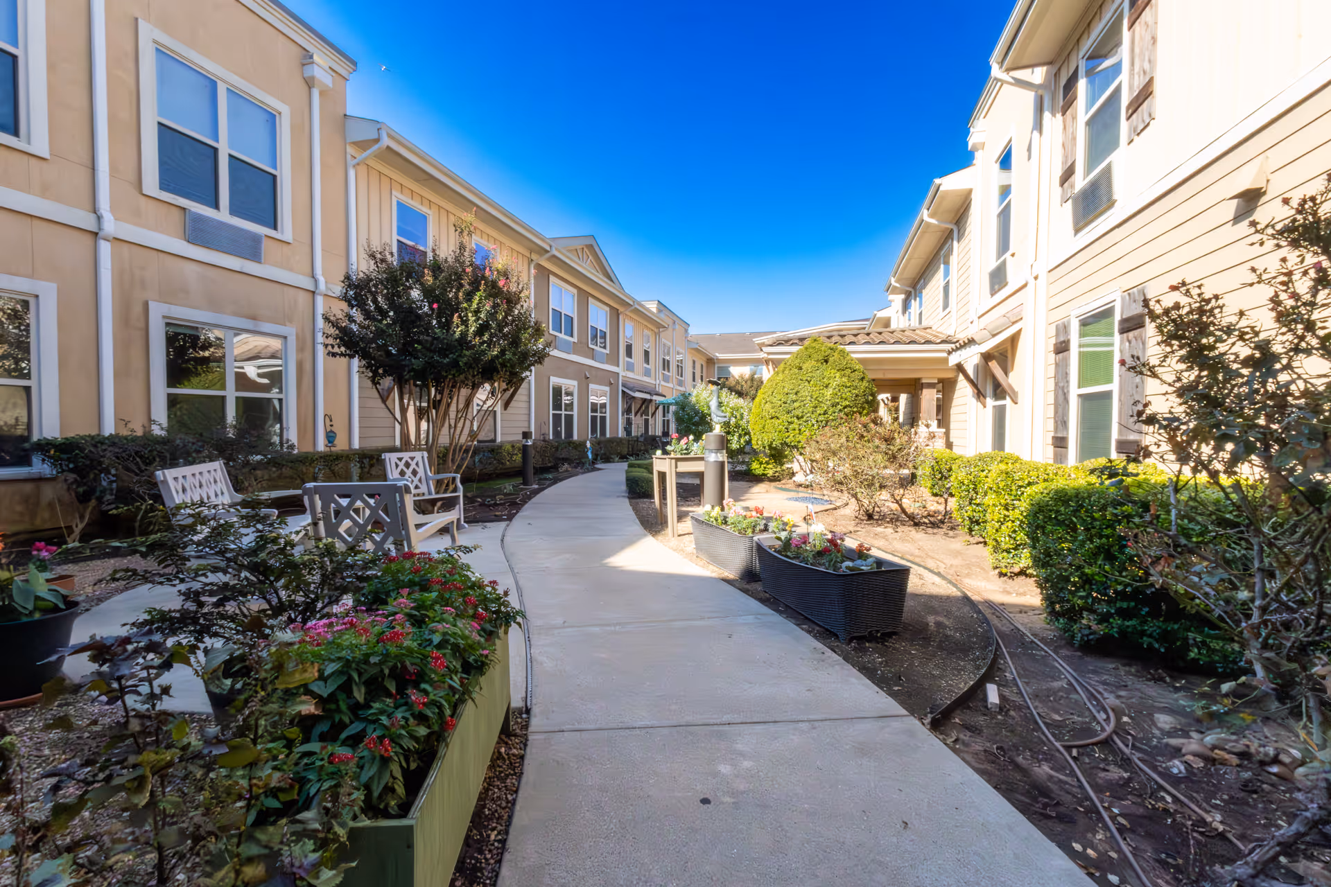 Outdoor courtyard area at Landon Ridge Sugar Land Assisted Living & Memory Care featuring a curved concrete walkway, flower planters, bushes, trees, and seating benches between two beige two-story buildings under a clear blue sky.