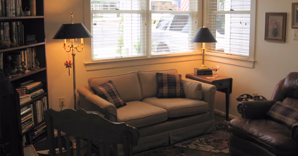 Cozy living room with a beige loveseat topped with plaid pillows, a bookshelf, lamps, and a leather armchair by a bay window.