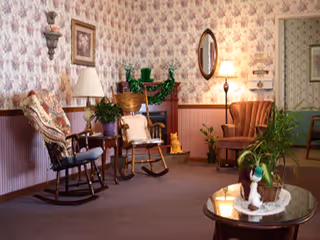 Cozy sitting area with rocking chairs, an upholstered armchair, a small fireplace, and plants on a coffee table in a wallpapered room.