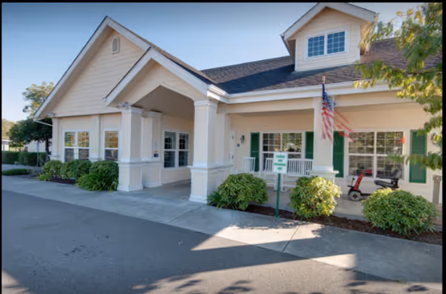 Exterior view of a single-story senior living community building with beige siding, white columns, green window shutters, and an American flag mounted near the entrance. There are bushes and a paved driveway in front of the building.