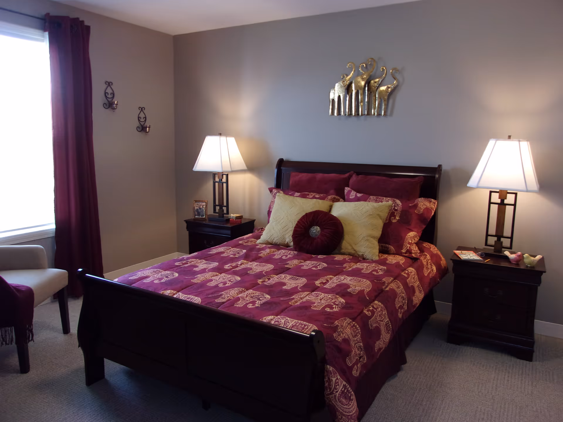 A cozy bedroom with a dark wooden bed frame and a bedspread featuring a maroon and gold elephant pattern. The bed is adorned with multiple pillows in maroon and gold. There are two matching dark wooden nightstands on either side of the bed, each with a modern table lamp. A window with maroon curtains is on the left side, and a beige armchair is partially visible. The walls are painted light gray and decorated with a metal wall art piece of elephants above the bed and two small candle holders on the left wall.