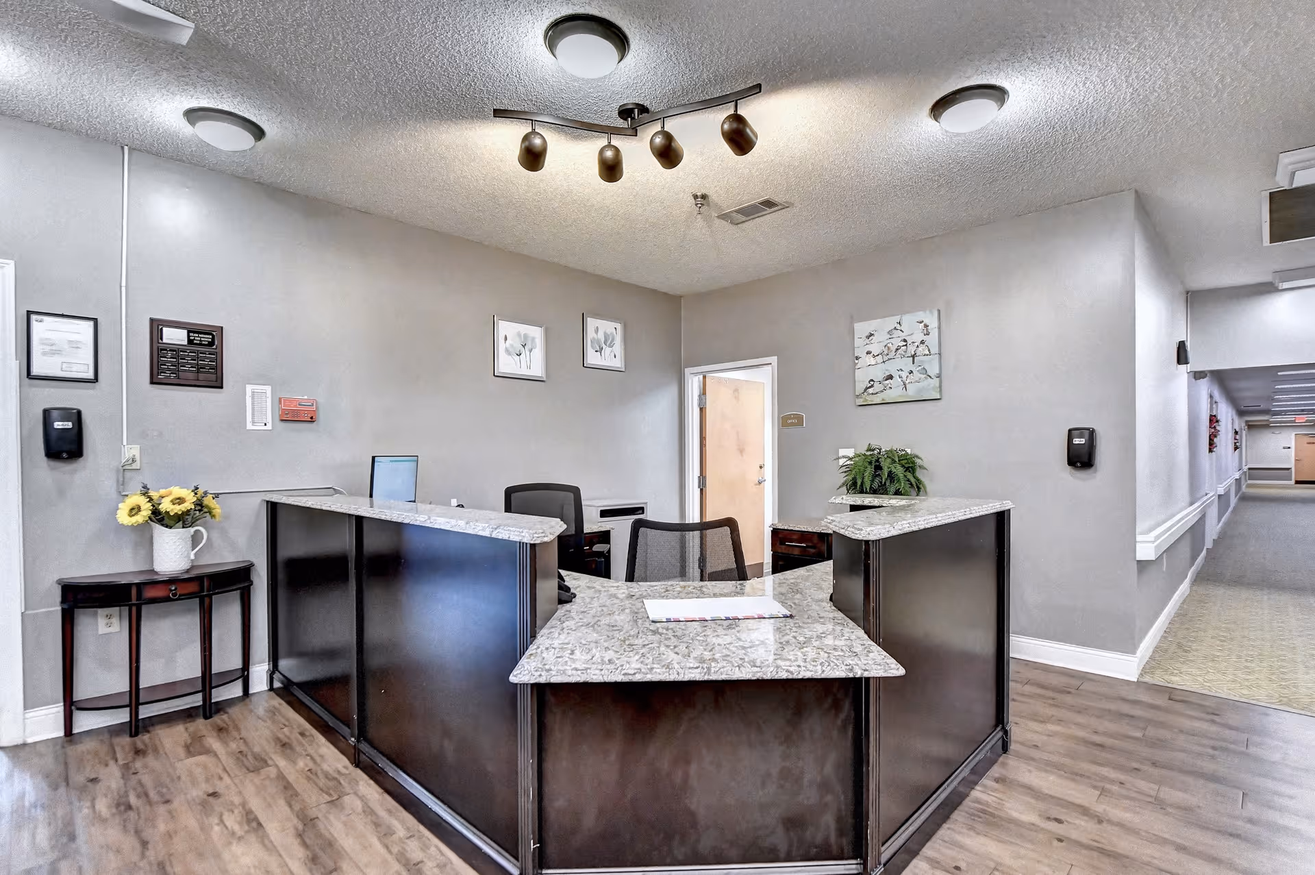 Reception desk and lobby of a senior living facility with a curved granite-topped counter, chairs, wall art, and a hallway.