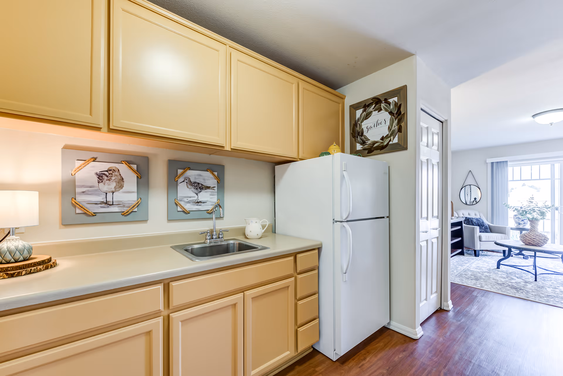 Small kitchen with beige cabinets, a sink, white refrigerator, decorative bird art, and a view into a bright living room.