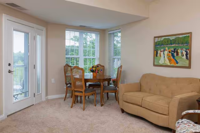Carpeted living area with a round dining table and chairs by bay windows, a beige loveseat, and a glass-paneled door to a balcony.
