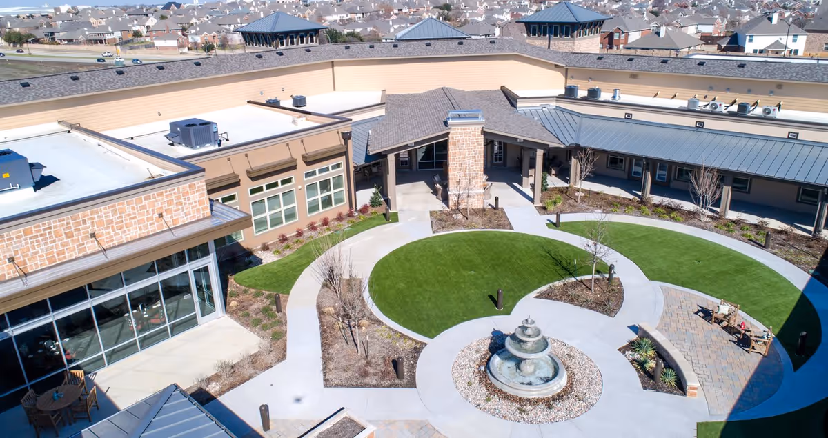 Aerial view of an outdoor courtyard at Discovery Village at Alliance Town Center Assisted Living, featuring a circular walkway surrounding a central fountain, green lawn areas, and seating spaces. The courtyard is enclosed by a single-story building with large windows and a covered entrance.