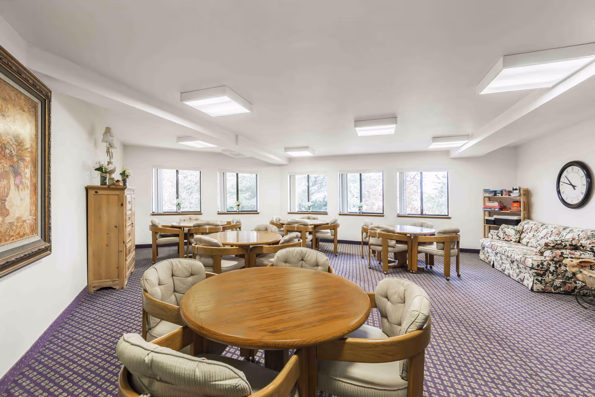 A bright room with multiple round wooden tables surrounded by cushioned chairs. The room has several windows letting in natural light, a floral-patterned couch against the wall, a large clock, a wooden cabinet, and a bookshelf with board games. The carpet is purple with a patterned design, and the walls are white.