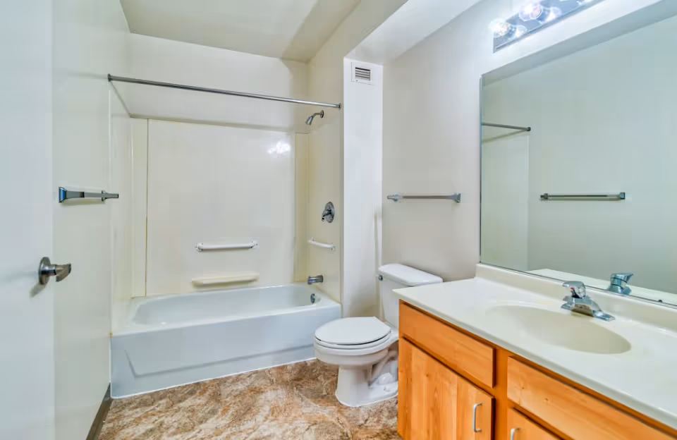 A bathroom with a bathtub/shower, toilet, and wooden vanity beneath a large mirror.