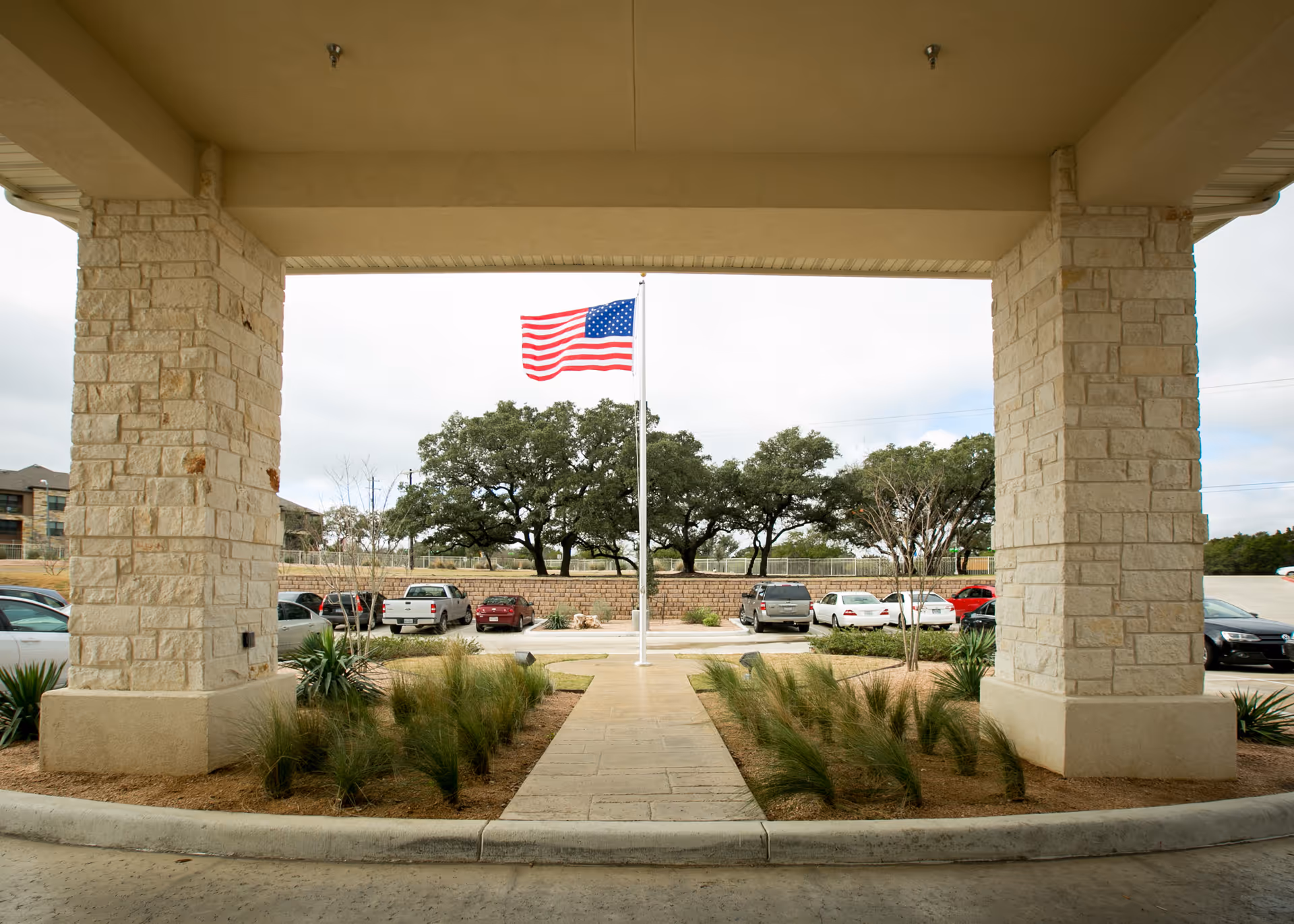 View from under a covered entrance with stone pillars looking out towards a parking lot, an American flag on a flagpole, and trees in the background.