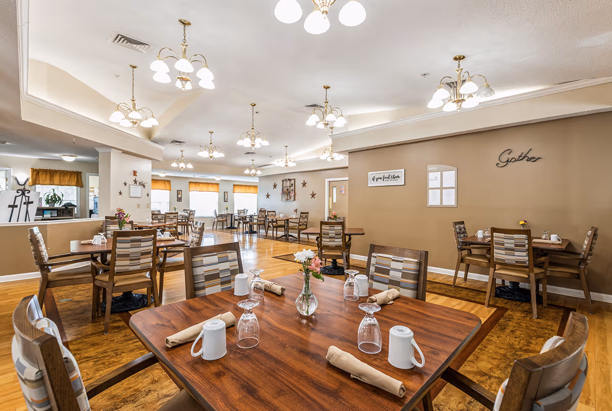 A spacious dining room with multiple wooden tables and chairs arranged neatly. Each table is set with white mugs, upside-down glasses, and beige napkins. The room has warm lighting from multiple ceiling chandeliers and large windows with orange valances letting in natural light. The walls are decorated with framed art and the word 'Gather' in cursive on one wall.