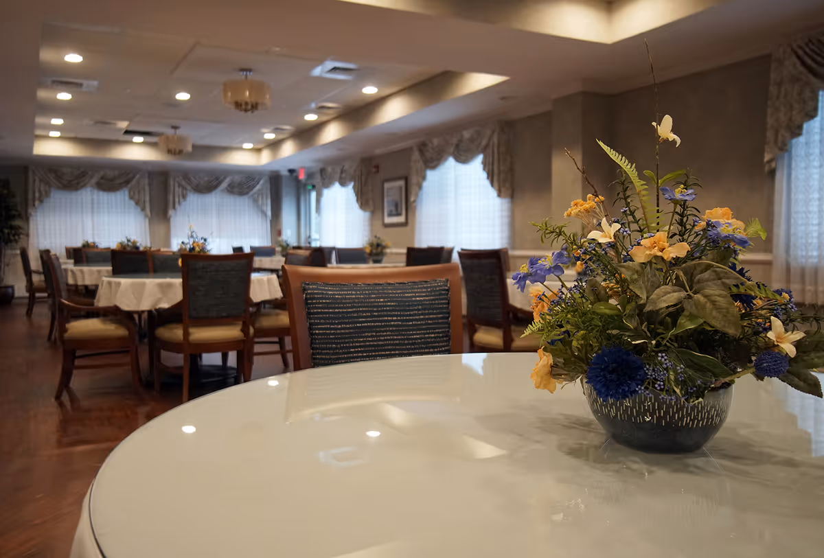 A dining room in a senior living facility with round tables covered in white tablecloths and wooden chairs with blue cushions. Each table has a floral centerpiece. The room has large windows with sheer curtains and valances, and ceiling lights provide warm illumination.