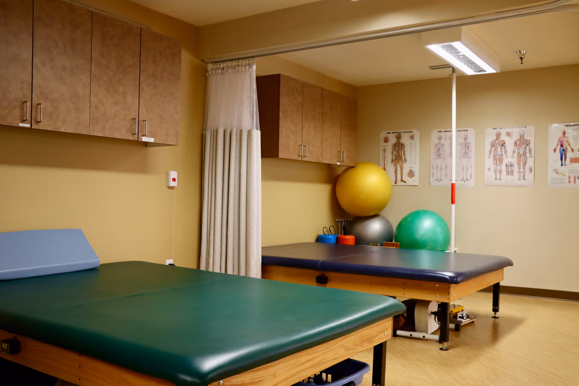 A therapy or rehabilitation room with two padded treatment tables, exercise balls in yellow, silver, and green, anatomical charts on the wall, beige walls, and wooden cabinets mounted above the tables.