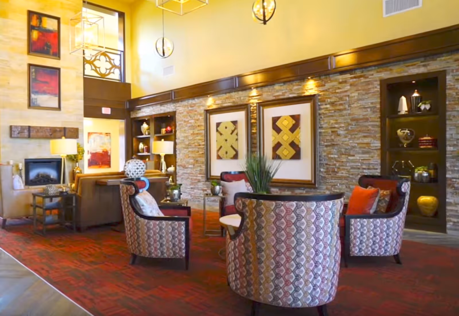 Stylish communal lounge with patterned armchairs and sofas arranged on a red carpet in front of a stone accent wall with artwork and warm lighting.