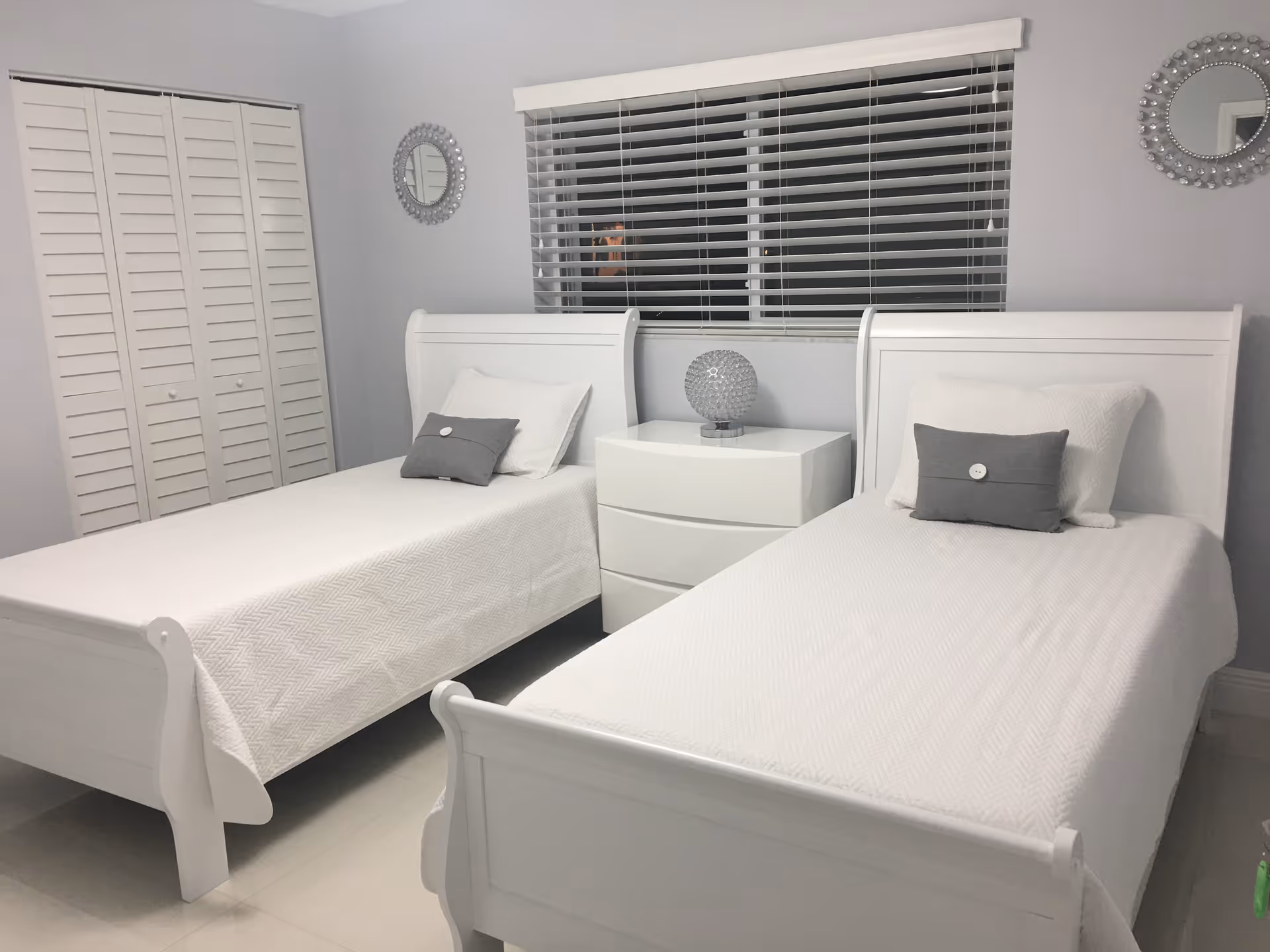 Minimalist bedroom with two white twin beds, a central white nightstand, and a window with blinds above it.