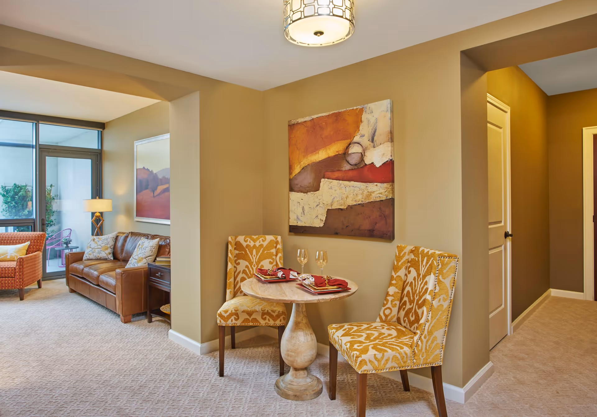 A cozy living area in a senior living facility featuring a small round table set for two with yellow patterned chairs, a brown leather sofa, an orange armchair, a floor lamp, and large windows leading to a balcony with outdoor seating. The walls are painted beige and decorated with abstract artwork.