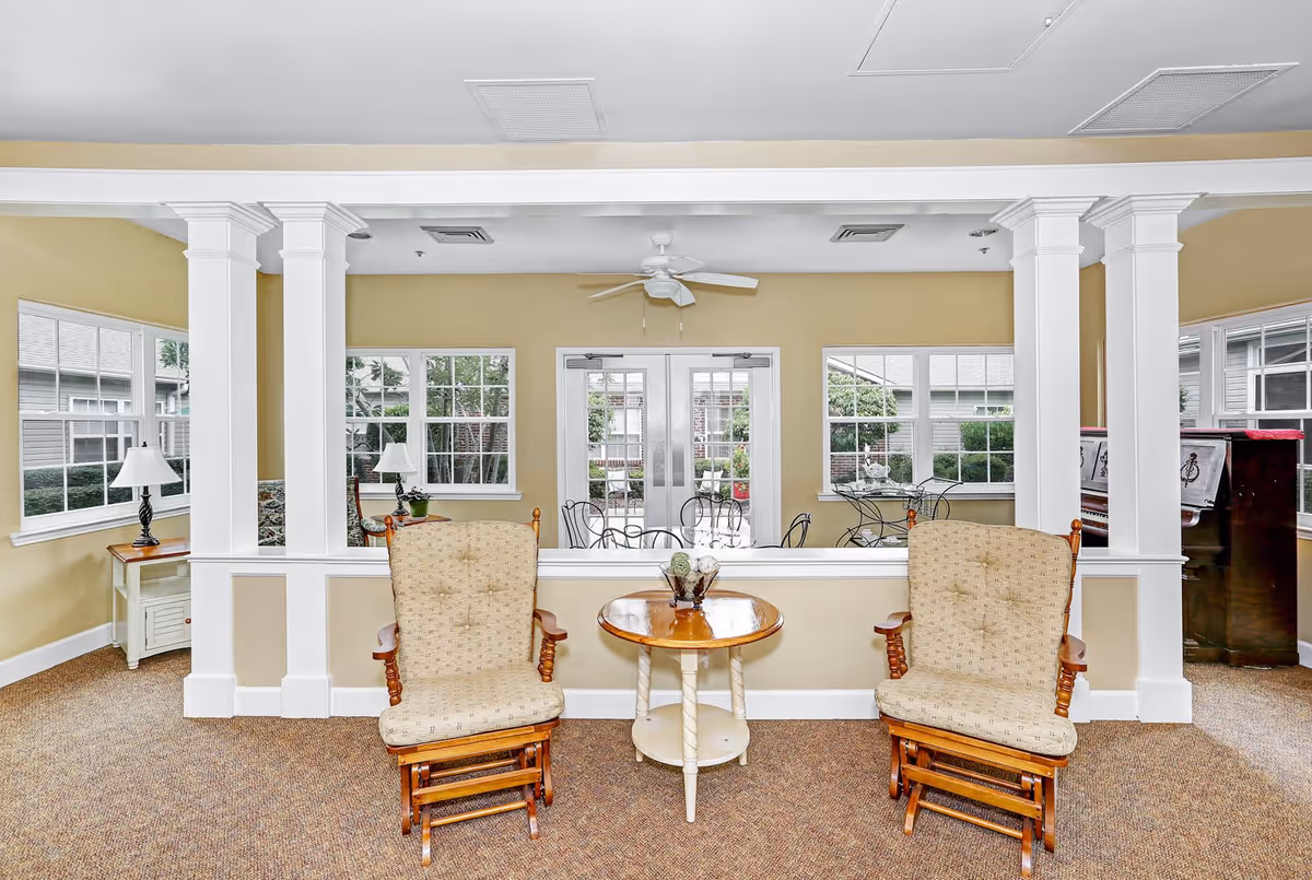 A cozy interior room with two cushioned wooden rocking chairs facing a small round wooden table with a decorative centerpiece. The room has beige walls, carpeted floor, white pillars, and large windows letting in natural light. Outside the windows, there is a patio area with metal chairs and tables. A ceiling fan is mounted above the door leading outside.