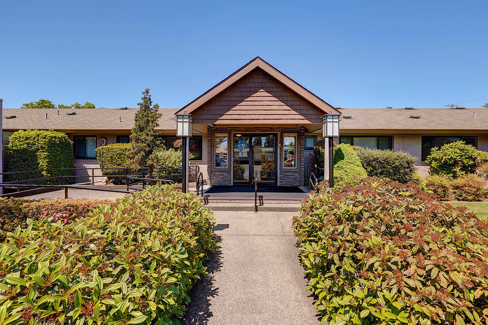 Front entrance of Gateway Care & Retirement Center with a paved walkway flanked by green bushes leading to double glass doors under a peaked roof. The building is single-story with a brown shingled roof and surrounded by well-maintained shrubs and plants under a clear blue sky.