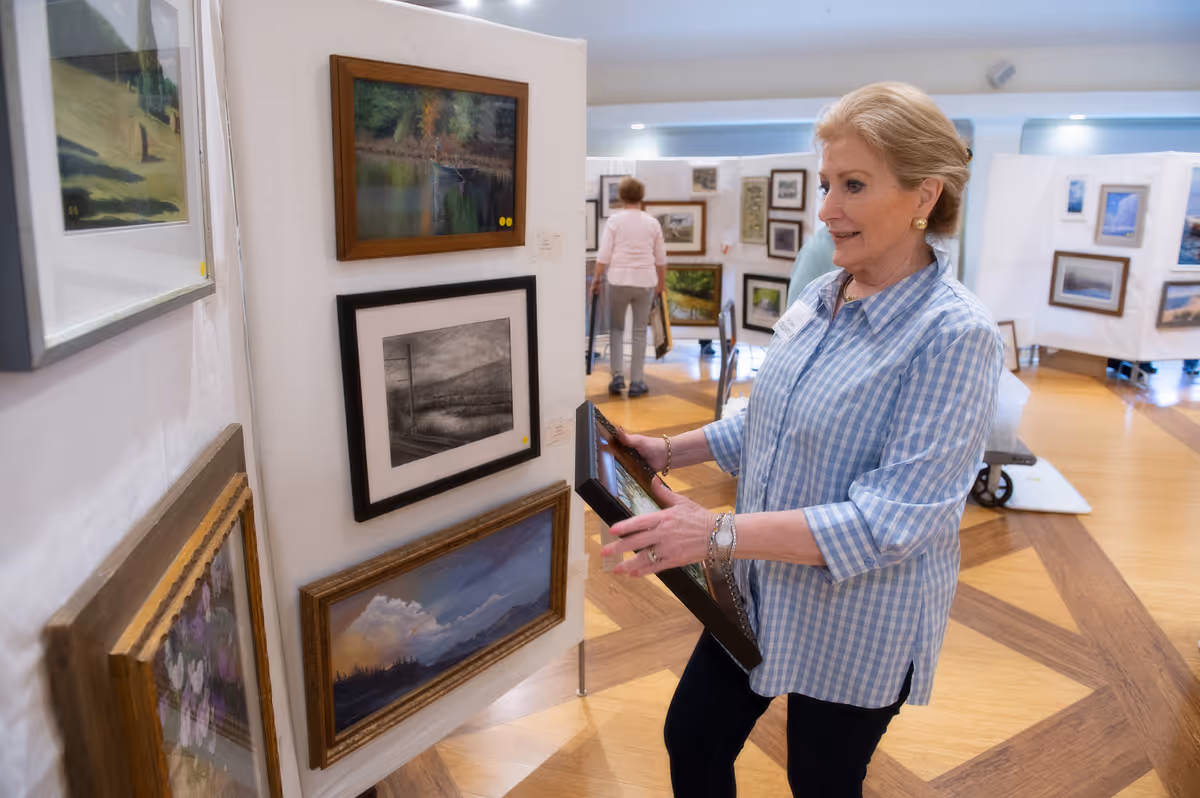 An elderly woman in a blue checkered shirt is viewing and holding framed artwork in an art gallery or exhibition space with several paintings displayed on white panels. Other people are also seen in the background looking at the artwork.