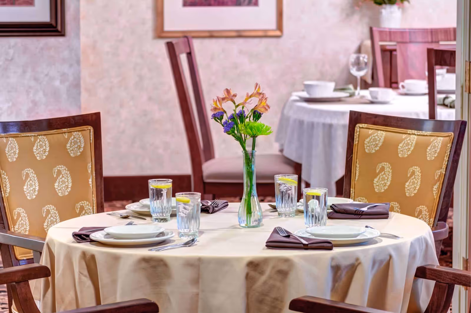 A round dining table set for four with beige tablecloth, white plates, silverware, glasses of water with lemon slices, and a vase with flowers in the center. The chairs have wooden frames with yellow patterned cushions. In the background, another similarly set table is visible.