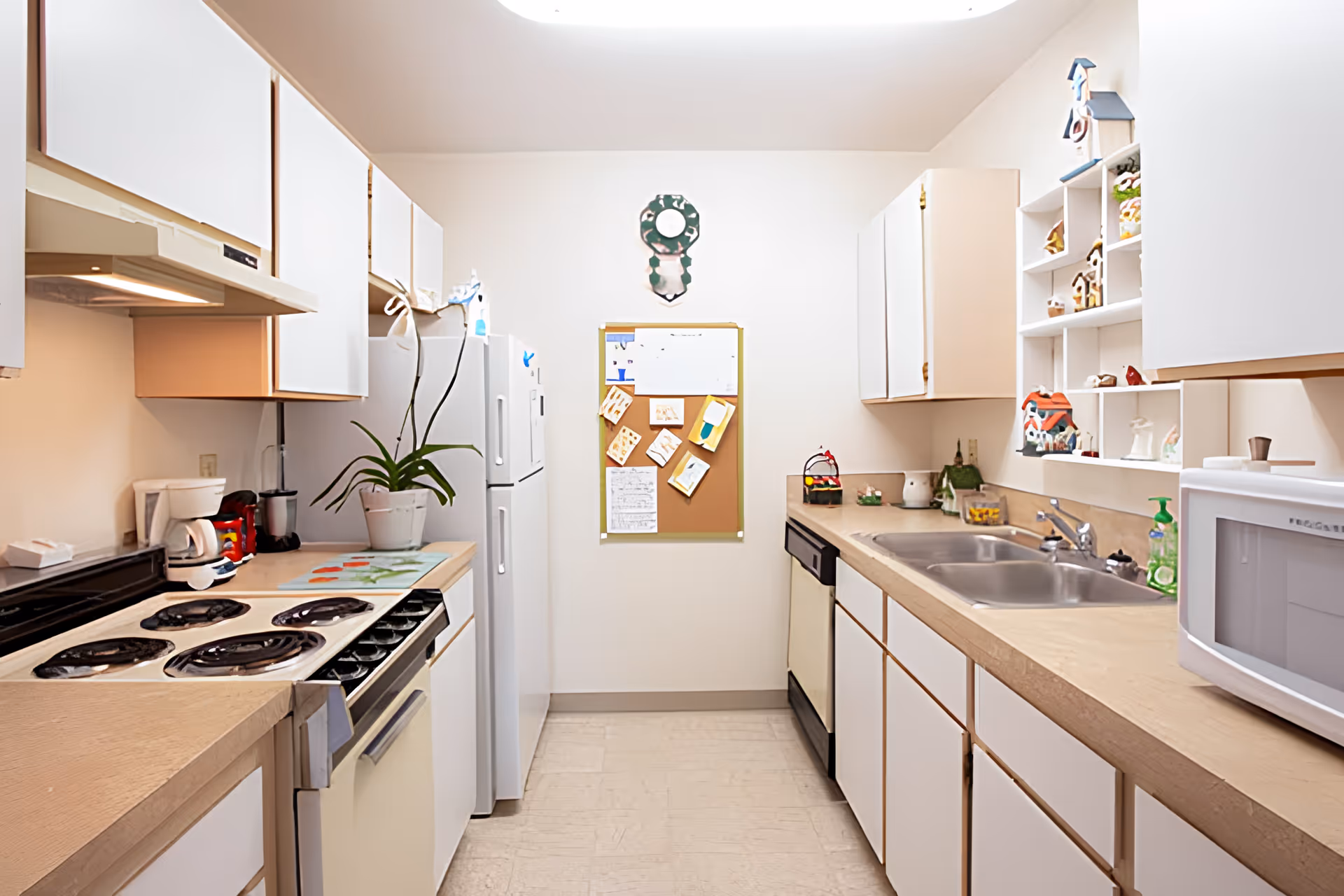 A bright kitchen with white cabinets and beige countertops. The kitchen features an electric stove, refrigerator, dishwasher, microwave, and a double sink. There are various small decorative items on the shelves and countertops, a potted plant, and a bulletin board with notes and pictures on the wall at the end of the kitchen.