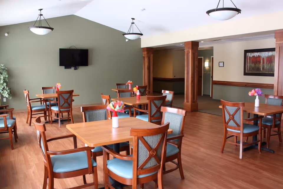 A dining room with several wooden tables and chairs with blue cushions. Each table has a small vase with colorful paper flowers. The room has wooden flooring, green walls, two hanging ceiling lights, a wall-mounted TV, and a framed painting on the wall. There are wooden columns and an open doorway leading to another area.
