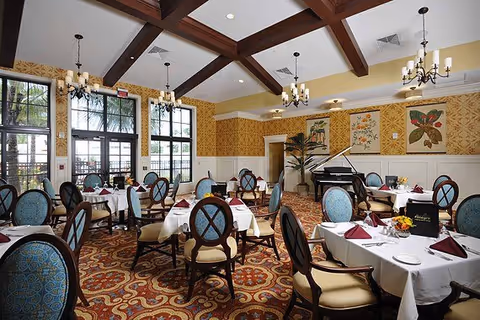 Elegant dining room with multiple round tables covered with white tablecloths and set with burgundy napkins, plates, and silverware. The room features high ceilings with wooden beams, chandeliers, large windows letting in natural light, patterned carpet, and a grand piano in the corner. Walls are decorated with framed botanical artwork and a tall plant near the piano.