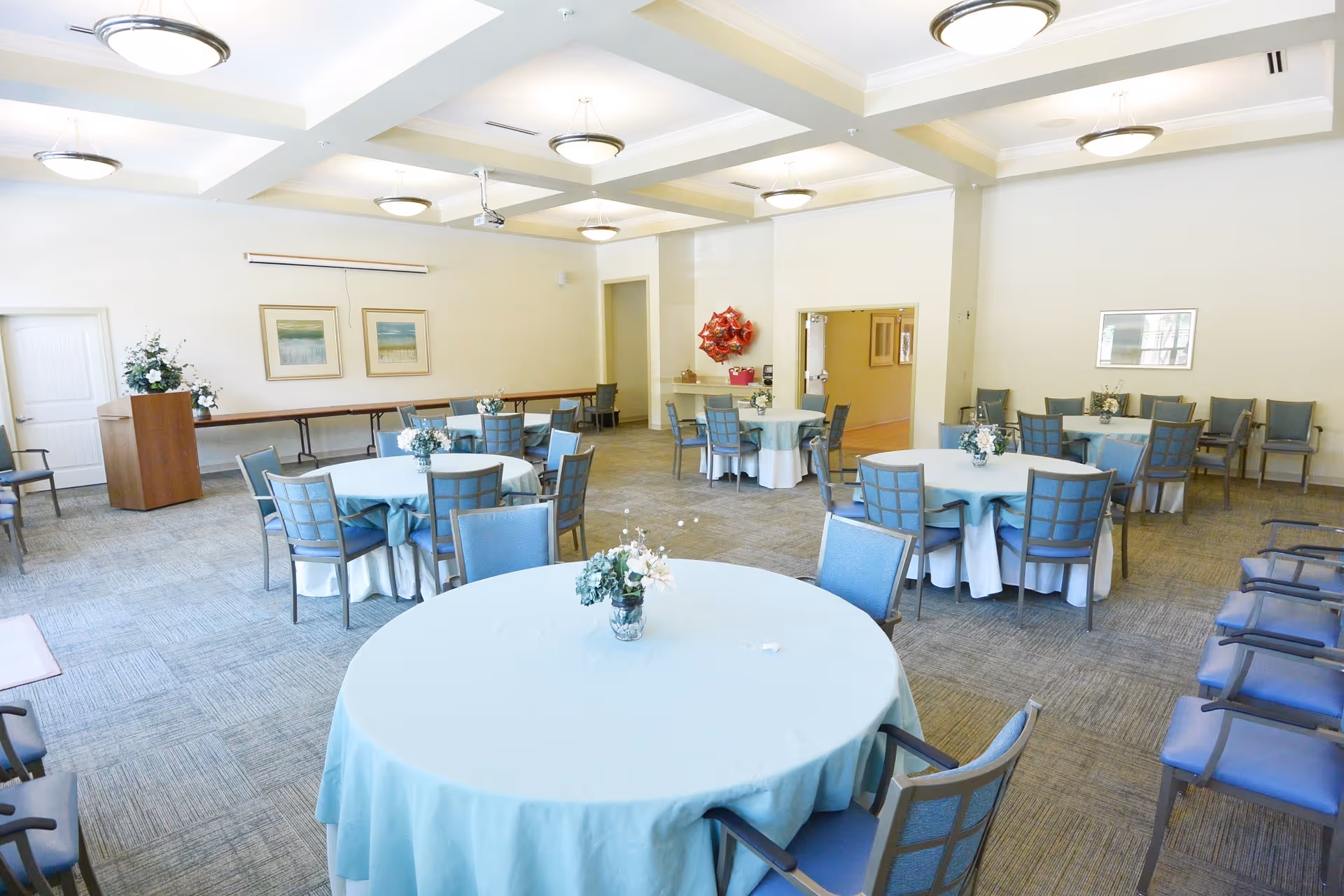 A bright and spacious dining room with round tables covered in light blue tablecloths, each adorned with a small floral centerpiece. The room has multiple chairs arranged around the tables and along the walls. The ceiling features recessed lighting and a projector. There are framed pictures on the walls and a door leading to another room.