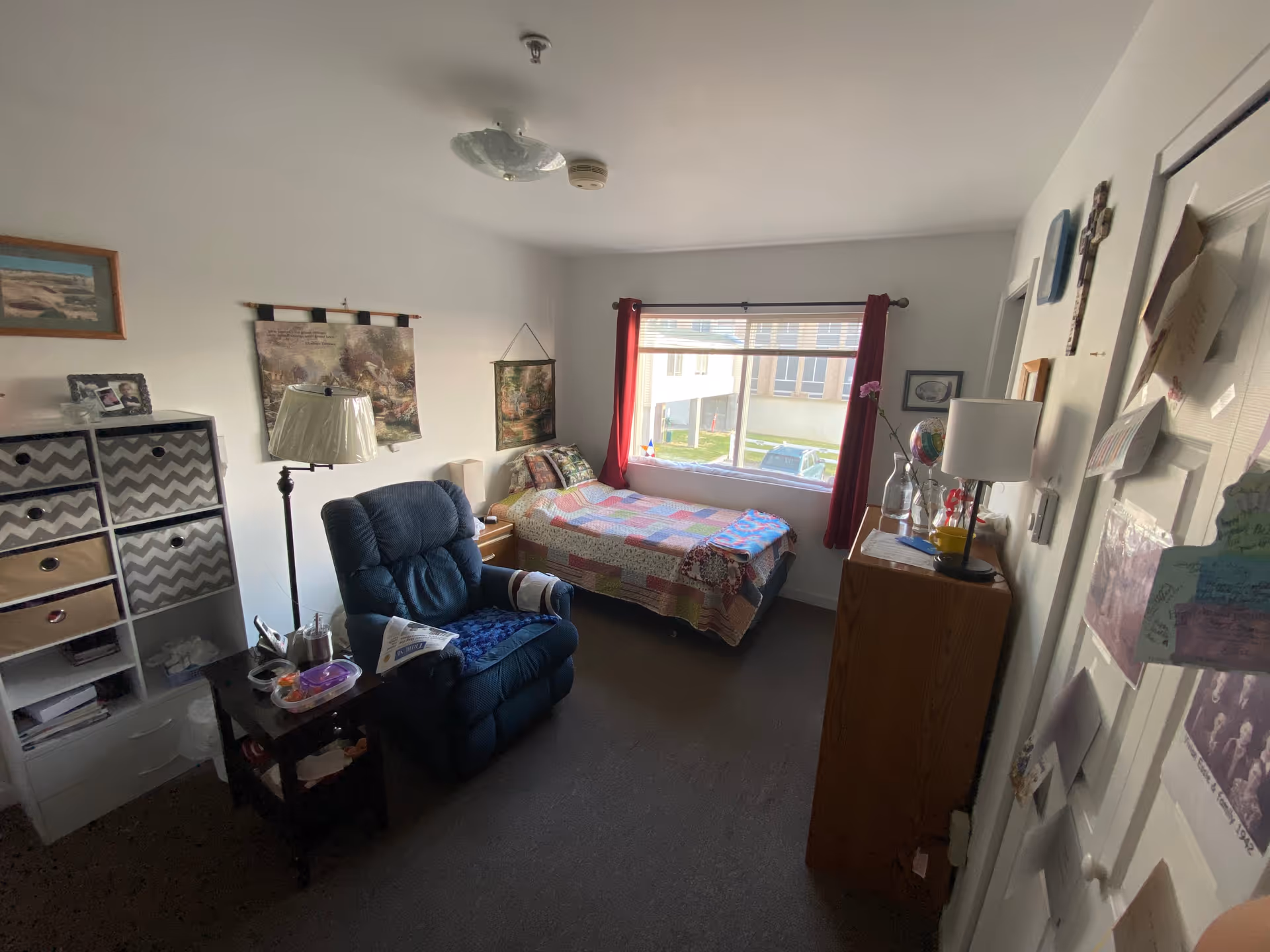 Small furnished bedroom with a single bed by a window, a recliner chair, dresser, and storage shelves.