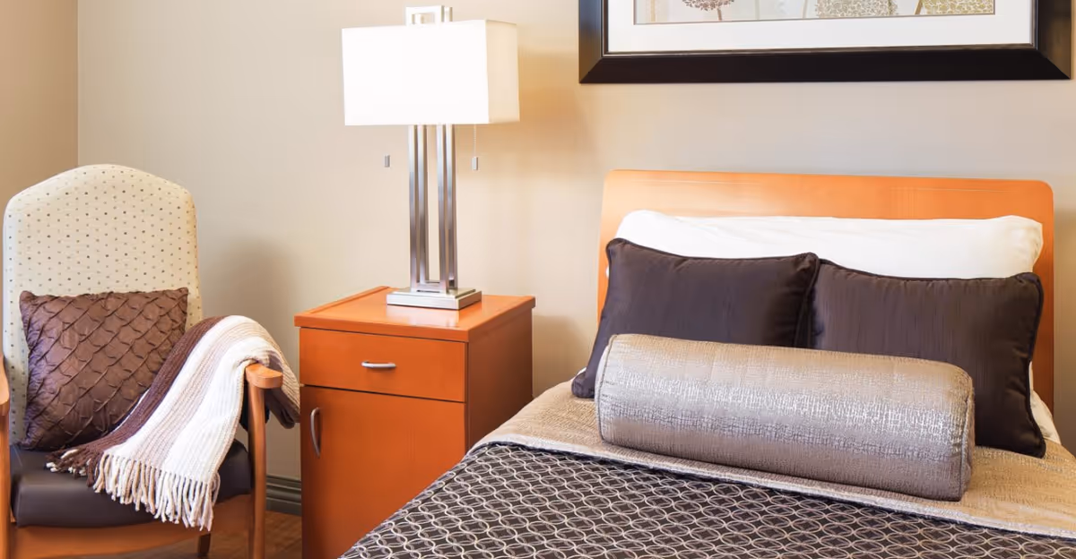 A cozy bedroom corner featuring a neatly made bed with multiple pillows including a large cylindrical bolster pillow, a wooden nightstand with a modern lamp, and an upholstered armchair with a textured pillow and a striped throw blanket draped over one arm.