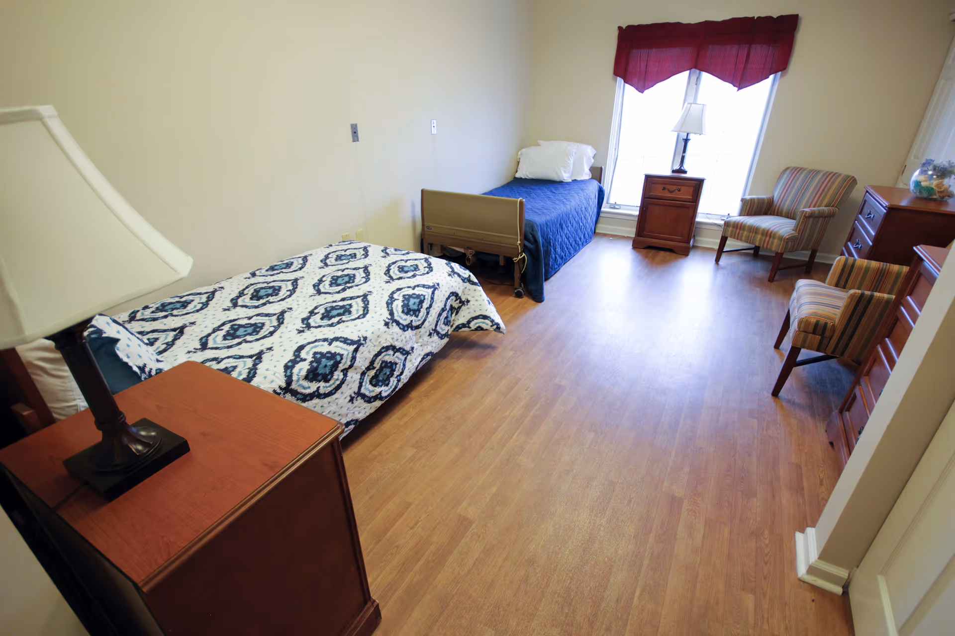A senior living facility bedroom with two single beds, one with a blue quilt and the other with a white and blue patterned quilt. The room has wooden flooring, a window with a red valance, a wooden nightstand with a lamp, two striped armchairs, and a wooden dresser with a decorative item on top.