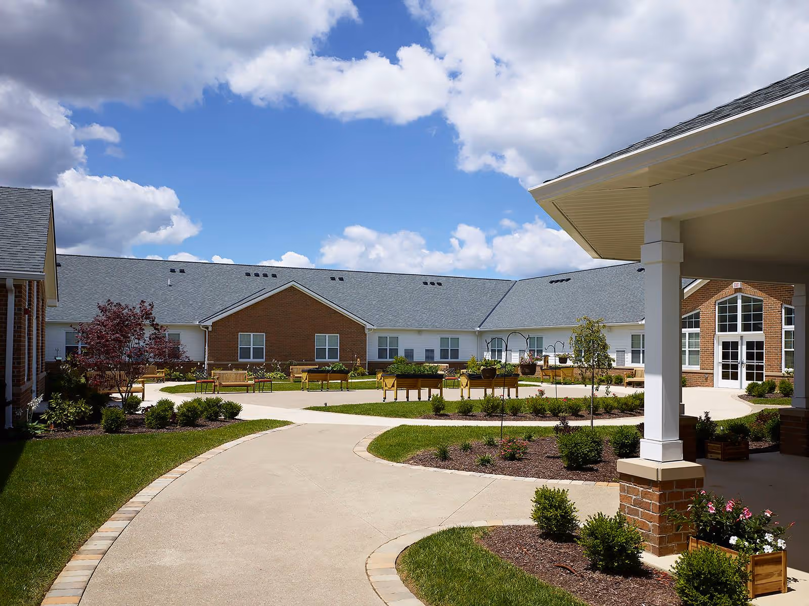 Sunny landscaped courtyard with walking paths, benches, and low brick-and-white buildings under a blue sky.