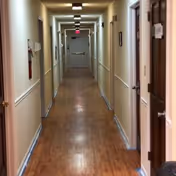 A long, narrow hallway with wooden flooring and beige walls in a senior living facility. Several doors line both sides of the hallway, and there are handrails along the walls. Ceiling lights illuminate the corridor, and an exit sign is visible above a door at the far end.