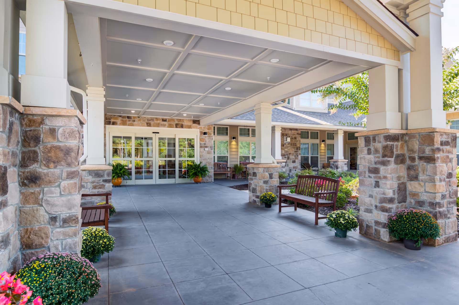 Covered entrance/portico of a senior living facility with stone columns, benches, potted plants, and double glass doors.