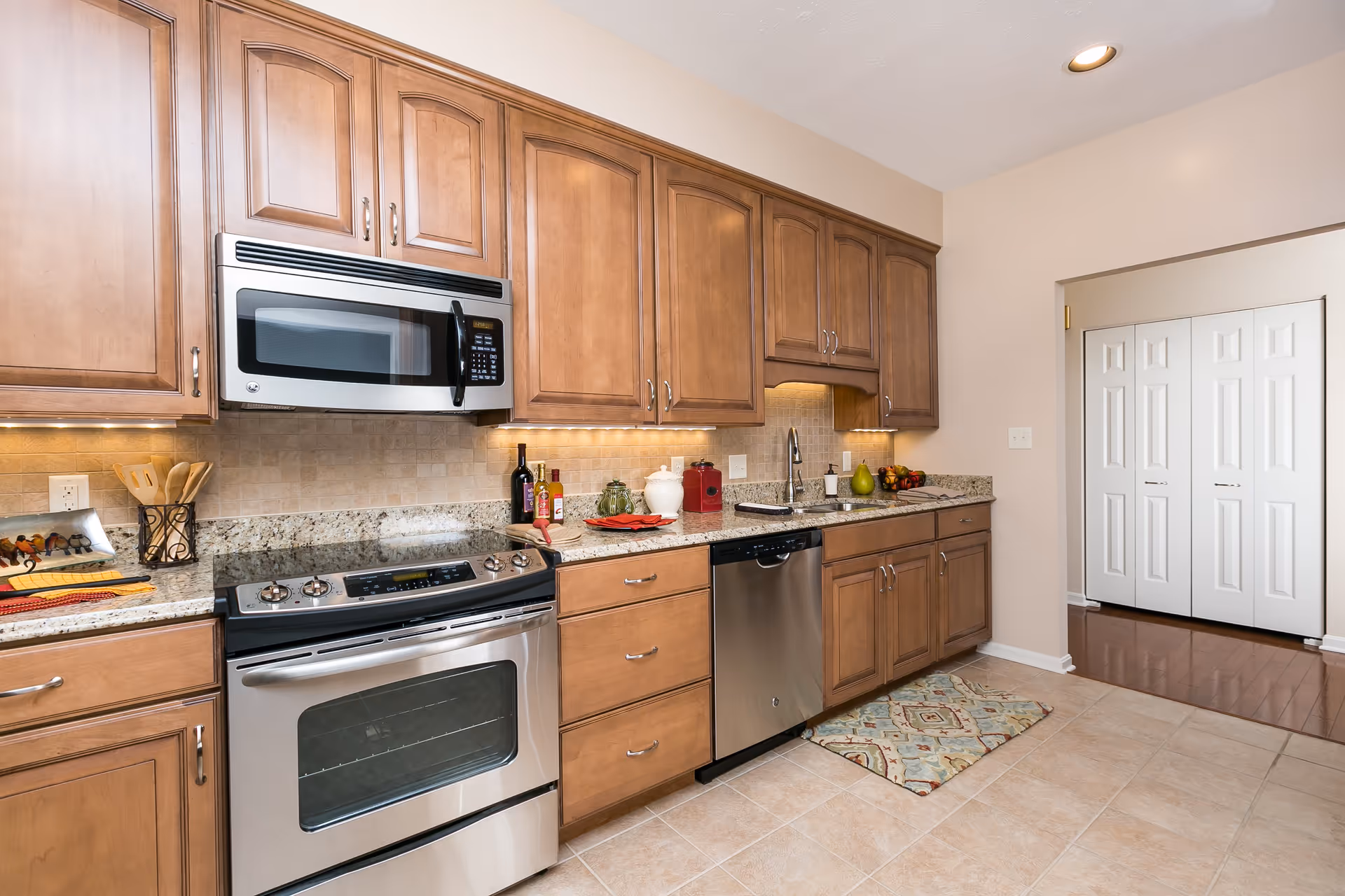 A modern kitchen with wooden cabinets, a stainless steel oven and microwave, a dishwasher, granite countertops, and a tiled floor. The kitchen is well-lit with under-cabinet lighting and recessed ceiling lights. There are various kitchen items on the countertop including bottles, utensils, and a fruit bowl. A patterned rug is placed on the floor in front of the sink area.