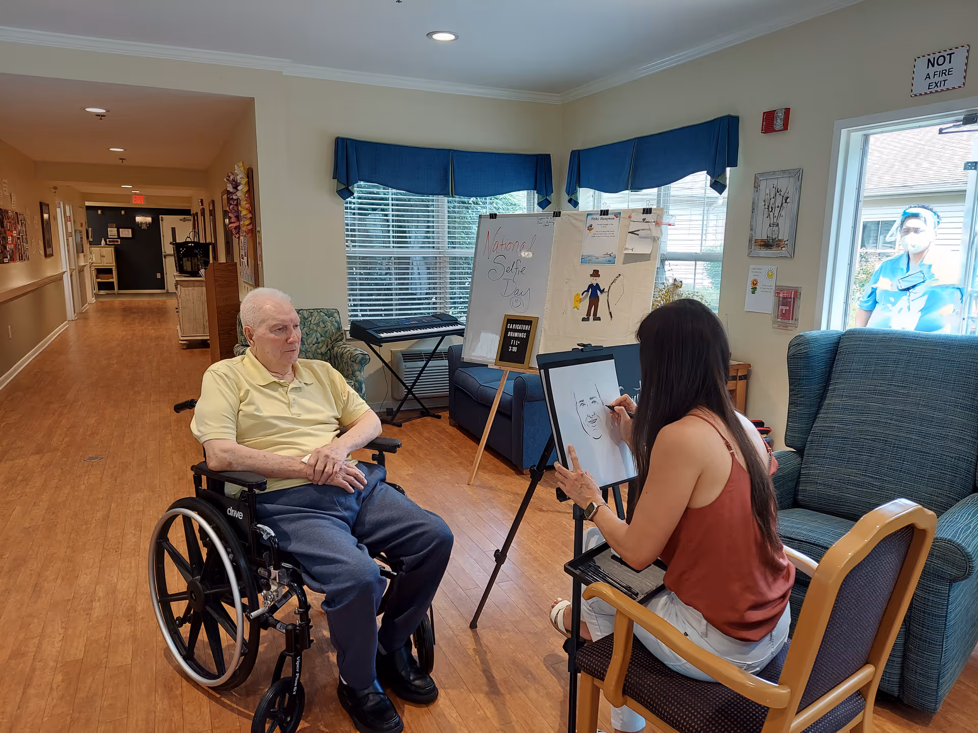 An elderly man in a wheelchair is sitting in a hallway of a senior living facility, while a woman seated in front of him is drawing his portrait on an easel. The room has wooden floors, a blue couch, a keyboard, and a whiteboard with notes and drawings. A person wearing a mask and blue scrubs is visible outside through a glass door.