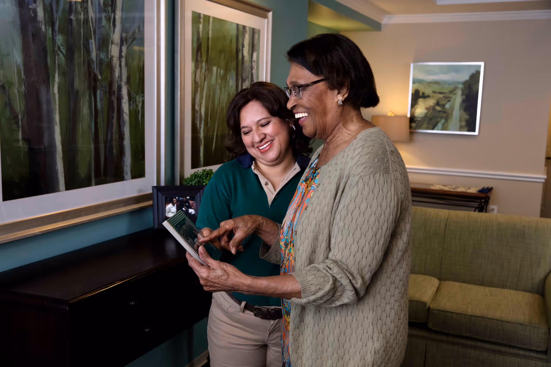 An elderly woman and a younger woman standing together in a living room area, smiling and looking at a photo album. The elderly woman is wearing glasses and a beige cardigan, while the younger woman is dressed in a green shirt and beige pants. Behind them is a green couch, framed artwork on the walls, and a dark wooden console table with a framed photo and a small plant.