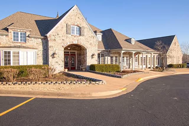 Exterior view of a stone-faced senior living facility entrance with an arched doorway, covered porch, and curved driveway.