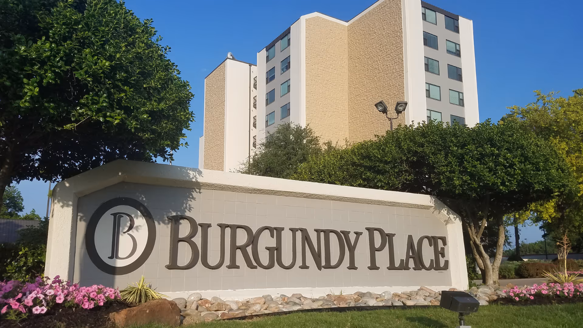 Outdoor view of the Burgundy Place senior living facility sign with a multi-story building and trees in the background under a clear blue sky.