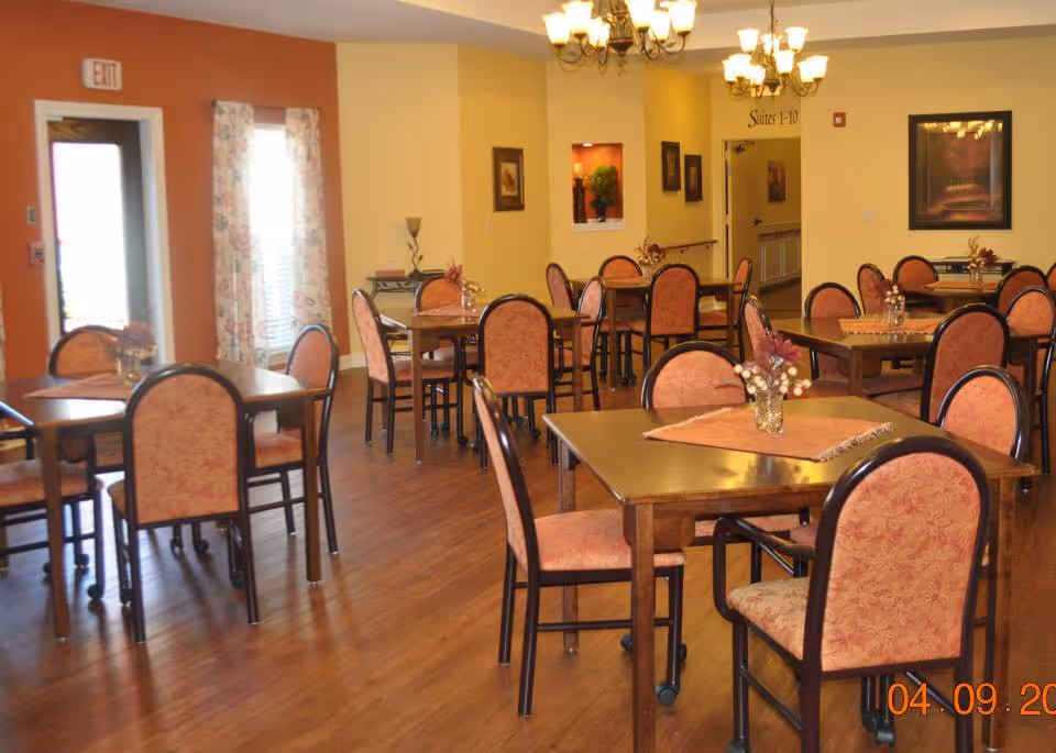 A dining room with multiple square and rectangular wooden tables surrounded by chairs with pink patterned upholstery. The room has warm yellow and orange walls, wooden flooring, and decorative floral curtains on the windows. There are small floral centerpieces on each table, and framed artwork on the walls. A chandelier with multiple lights hangs from the ceiling.