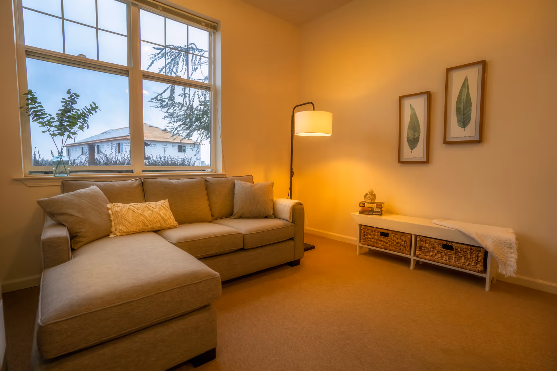 A cozy living room with a beige sectional sofa adorned with cushions, a floor lamp emitting warm light, two framed leaf prints on the wall, and a white bench with wicker baskets underneath. A window shows a view of a house under construction and some greenery outside.