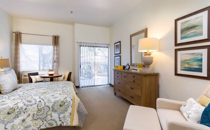 A bright and cozy bedroom featuring a bed with patterned bedding, a wooden dresser with a lamp and mirror above it, a comfortable armchair with a pillow, and a small round table with two chairs near a window. Vertical blinds cover a sliding glass door leading outside.