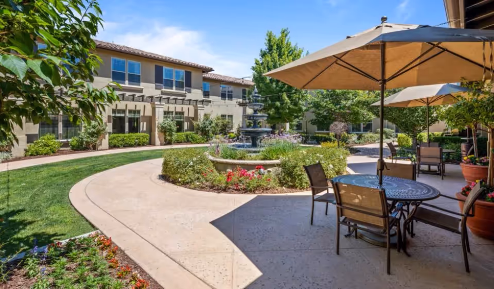 Outdoor courtyard area at Oakmont of Fresno featuring a circular fountain surrounded by greenery and flowers, with patio tables and chairs under large umbrellas on a sunny day.