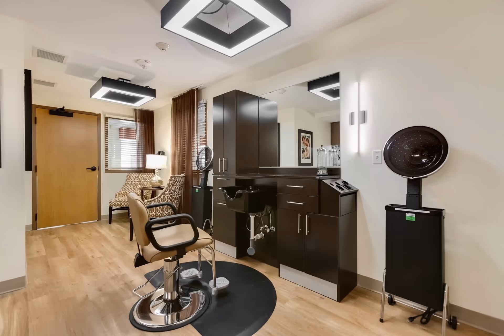 Interior view of a hair salon area in a senior living facility with a salon chair, a black sink, dark wood cabinets, a large mirror, two patterned chairs near a window with brown curtains, and a hair dryer on a stand against a white wall.