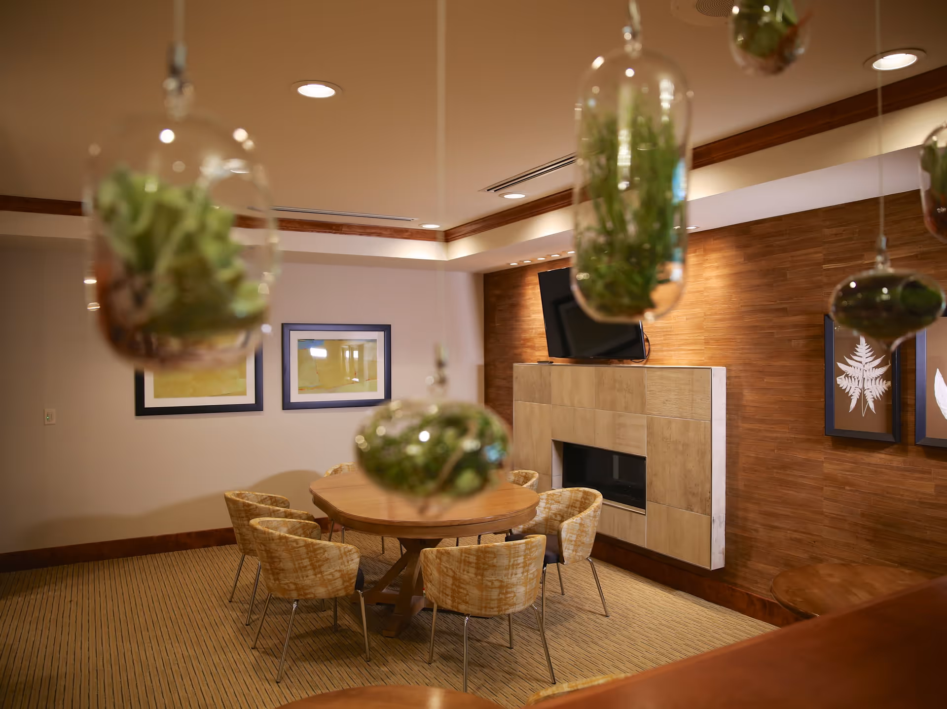 A cozy interior room with a round wooden table surrounded by six cushioned chairs. The room features a wall-mounted flat-screen TV above a modern fireplace with beige tiles. The walls are decorated with framed artwork and wooden paneling. Several glass terrariums with green plants hang from the ceiling in the foreground.