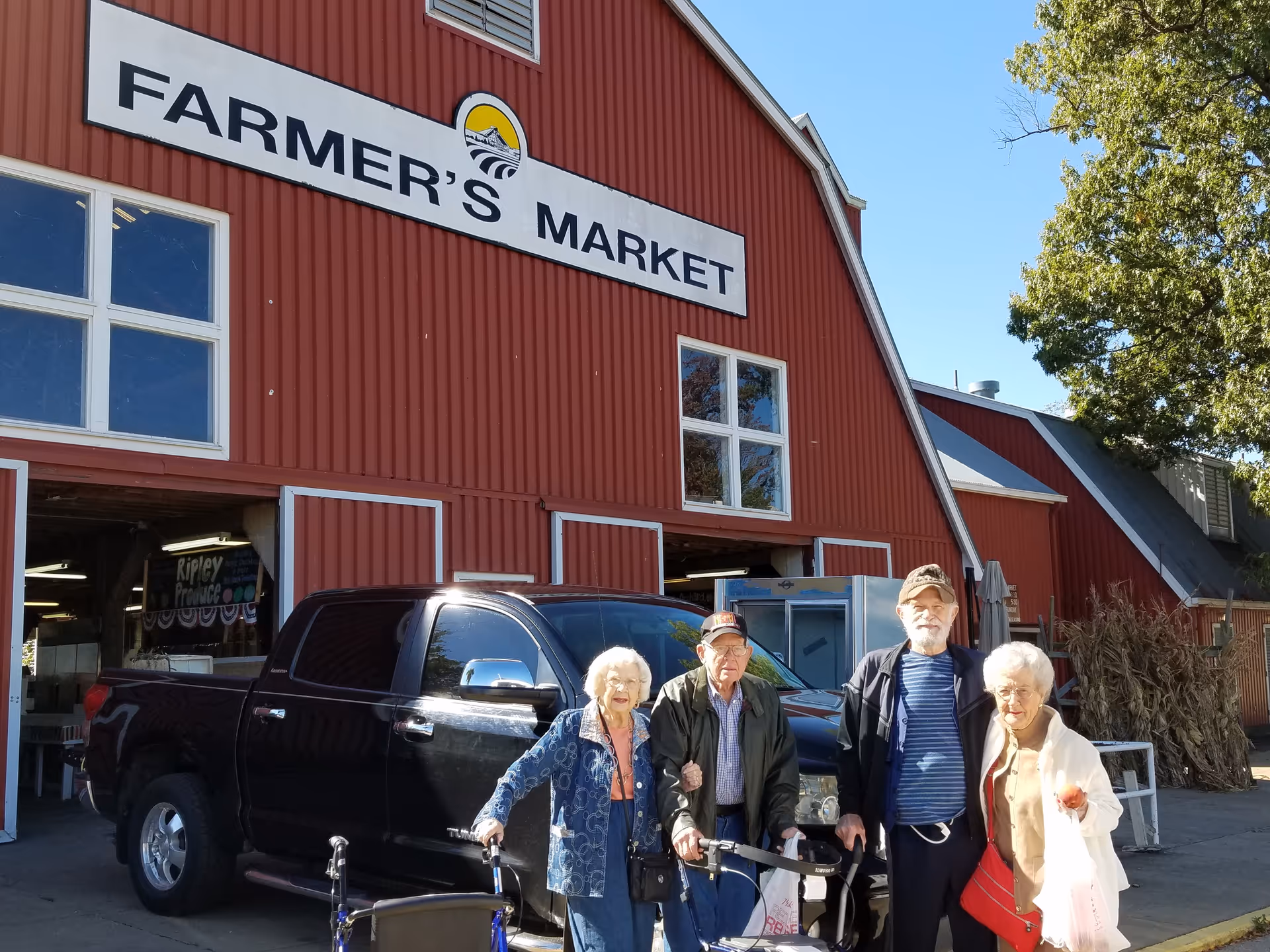Four elderly people standing outside a red building with a sign that reads 'FARMER'S MARKET'. There is a black pickup truck parked behind them and a tree to the right.
