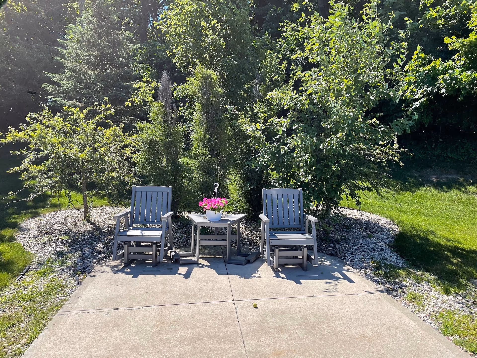 Two gray wooden chairs with armrests and a small matching table between them on a concrete patio. On the table is a white pot with pink flowers. Behind the chairs are green bushes and trees with a grassy area surrounding the patio.