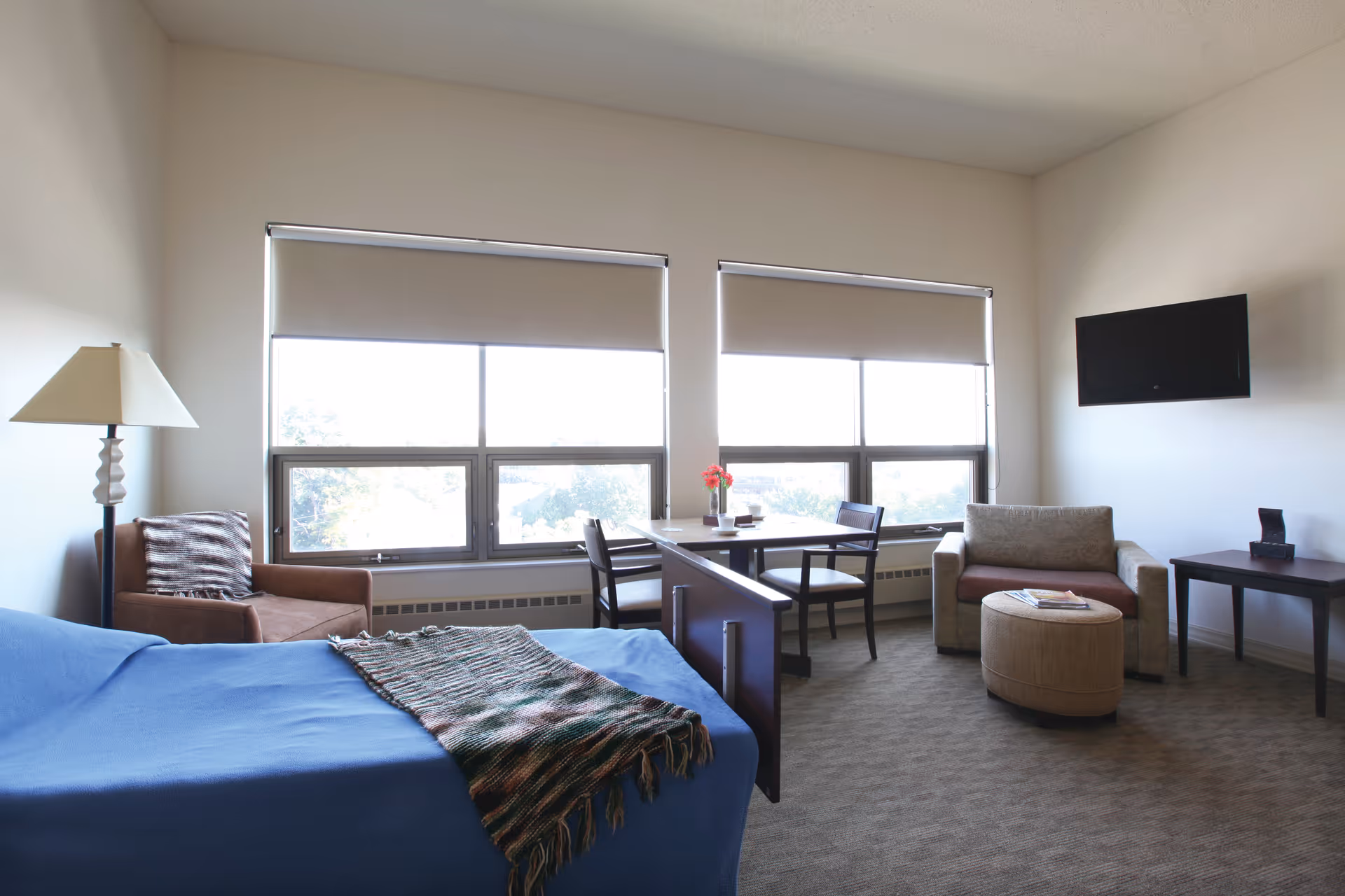 A bright and spacious room with a bed covered in a blue blanket and a patterned throw, a brown armchair with a striped pillow, a small dining table with two chairs near large windows with blinds, a beige loveseat, a round ottoman, a side table, and a wall-mounted flat-screen TV.