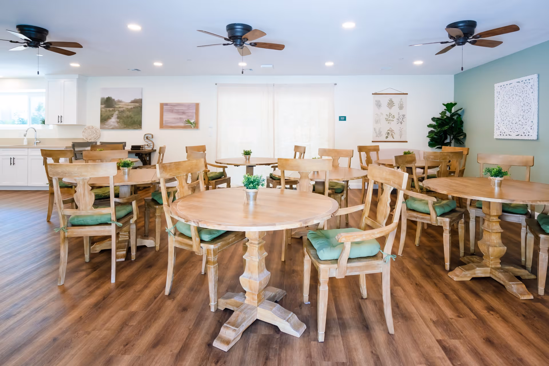 A bright and spacious dining room with multiple round wooden tables and matching wooden chairs with green cushions. The room has wood flooring, ceiling fans, and light-colored walls decorated with framed artwork and plants. There is a kitchen area visible in the background with white cabinets and a sink.